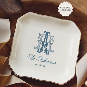 May include: A square white ceramic trinket dish with scalloped edges, featuring a blue monogram of the letters A and H, the text The Andersons, and the date 05.18.2027. A white scalloped label in the corner displays the text GIFT BOX INCLUDED.