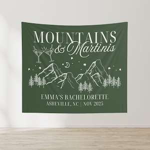 May include: Green fabric banner with white text and illustrations. The text reads "Mountains & Martinis" and "Emma's Bachelorette Asheville, NC | Nov 2025". The illustration depicts mountains, trees, stars, a crescent moon, and two martini glasses.