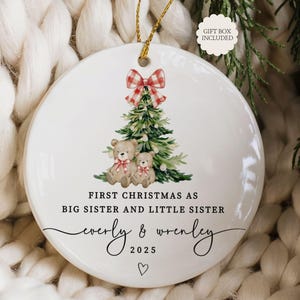 May include: A white ceramic Christmas ornament with a watercolor design. It features a Christmas tree, two teddy bears, and a red and white plaid bow. Text reads "First Christmas as Big Sister and Little Sister" with names and the year 2025.