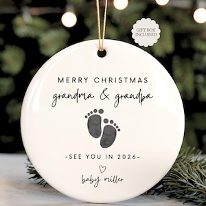 May include: A white ceramic Christmas ornament with the text "MERRY CHRISTMAS grandma & grandpa" and baby footprints. The ornament also includes the text "- SEE YOU IN 2026 -" and "baby miller". A gift box is included.