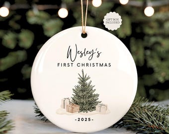 May include: A white ceramic Christmas ornament with the text "Wesley's FIRST CHRISTMAS - 2025". The ornament features a watercolor illustration of a Christmas tree and wrapped gifts. A gold string is attached for hanging.