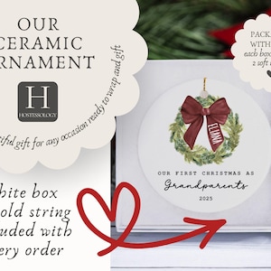 May include: A round white ceramic ornament featuring a watercolor green wreath with a deep red ribbon printed with the name Eliana. The text reads Our First Christmas as Grandparents 2025. The ornament is displayed inside a square white gift box.