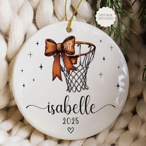 May include: White ceramic basketball ornament with a brown bow and basketball hoop design. The name "Isabelle" and the year "2025" are written in black script. Includes a gold string for hanging. Gift box included.