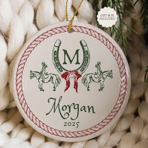 May include: A white ceramic ornament with a red rope border and gold hanging cord. The ornament features a horseshoe, riders on horses, and the name "Morgan" with the year "2025" in green script. A red bow is below the horseshoe.