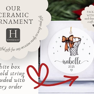 May include: White ceramic ornament with a basketball hoop design, a brown bow, and the name "Isabelle" with the year 2025. The ornament is packaged in a white box with a gold string. Text on the ornament reads "Our Ceramic Ornament" and "a beautiful gift for any occasion ready to wrap and gift."