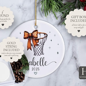 May include: White ceramic basketball ornament, 3 inches in diameter, with a basketball hoop design and an orange bow. Includes a gold string for hanging. Features the name "Abelle" and the year 2025.