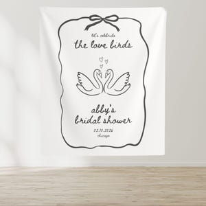 May include: White bridal shower backdrop with a black border and a graphic of two swans with hearts. The text reads "Let's celebrate the love birds Abby's bridal shower 02.10.2026 Chicago"