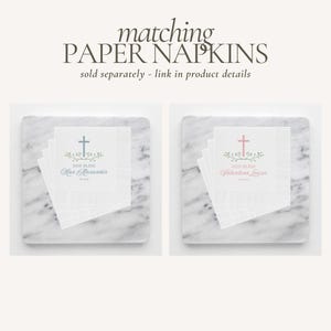 May include: Two white paper napkins with a cross and floral design. The text "God Bless" is printed on each napkin, along with a name. One napkin has a blue cross and the name "Max Alexander". The other napkin has a pink cross and the name "Valentina Louise".