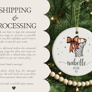 May include: A white ceramic Christmas ornament with a basketball hoop design and a decorative orange bow. The ornament includes the name "isabelle", the year "2025", and a heart symbol. It hangs from a gold string.
