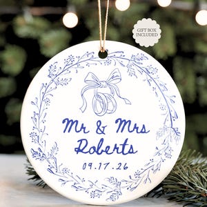 May include: A white ceramic ornament with a blue floral design and the text "Mr & Mrs Roberts 09.17.26". The ornament features a bow and two intertwined rings. A gift box is included.