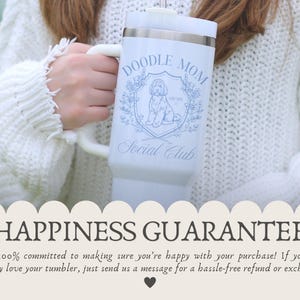 May include: A white tumbler with a handle, featuring the text "DOODLE MOM Social Club" and a doodle dog illustration in blue. The tumbler is held by a person wearing a white sweater. The bottom of the image has the text "HAPPINESS GUARANTEE".