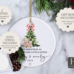 May include: A white ceramic Christmas ornament, 3 inches in diameter, with a gold string. It features a Christmas tree design with teddy bears and the text "First Christmas as Sister and Little Sister" with a name and the year 2025.