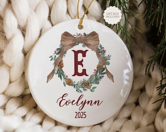 May include: A white ceramic Christmas ornament with a brown bow and a wreath design. The ornament features the letter "E" in burgundy, the name "Evelynn", and the year "2025". A gold string is attached for hanging.