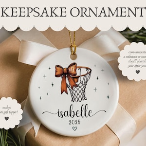 May include: White ceramic keepsake ornament with a basketball hoop design and a brown bow. The ornament has the name "Isabelle" and the year "2025" written on it. The text "KEEPSAKE ORNAMENT" is at the top.