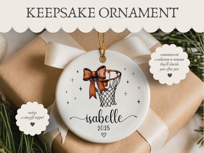 May include: White ceramic keepsake ornament with a basketball hoop design and a brown bow. The ornament has the name "Isabelle" and the year "2025" written on it. The text "KEEPSAKE ORNAMENT" is at the top.