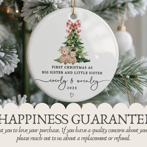 May include: White ceramic Christmas ornament with a festive design. It features a Christmas tree, teddy bears, and a red and white plaid bow. Text reads "First Christmas as Big Sister and Little Sister" with names and the year 2025.