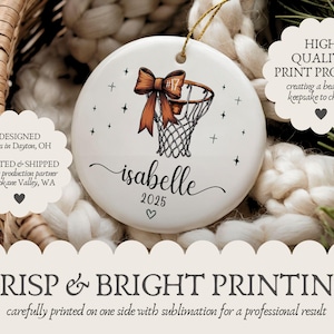 May include: White ceramic ornament featuring a basketball hoop with an orange bow. The ornament includes the name "isabelle" and the year "2025". Black star accents surround the basketball design. A gold string is attached for hanging.