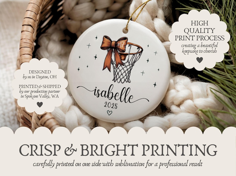 May include: White ceramic ornament featuring a basketball hoop with an orange bow. The ornament includes the name "isabelle" and the year "2025". Black star accents surround the basketball design. A gold string is attached for hanging.