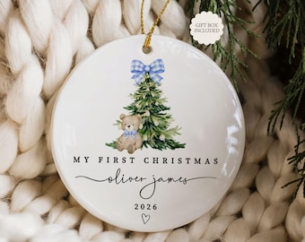 May include: A round white ceramic ornament featuring a watercolor illustration of a green Christmas tree with a blue gingham bow and a teddy bear wearing a blue gingham bowtie. The text reads MY FIRST CHRISTMAS, oliver james, and 2026.