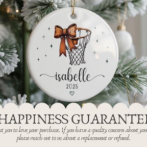 May include: A white ceramic Christmas ornament featuring a basketball hoop design with a brown bow and the name "isabelle" in cursive. The year "2025" and a heart are also included. The ornament is hanging on a Christmas tree.
