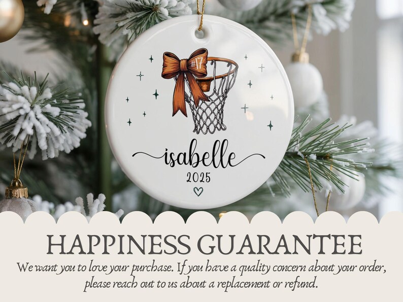 May include: A white ceramic Christmas ornament featuring a basketball hoop design with a brown bow and the name "isabelle" in cursive. The year "2025" and a heart are also included. The ornament is hanging on a Christmas tree.