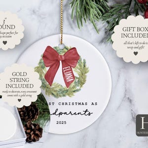 May include: A 3-inch round white ceramic ornament featuring a watercolor green wreath with a red bow and the name ELIANA printed on the ribbon. The text reads FIRST CHRISTMAS AS grandparents 2025. It includes a gold string and a gift box.