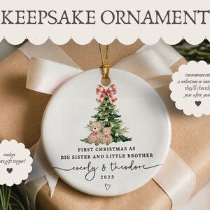 May include: A white ceramic Christmas ornament with a gold string, featuring a Christmas tree and two teddy bears. The ornament reads "First Christmas as Big Sister and Little Brother" with names and the year 2025. A red and white plaid bow tops the tree.