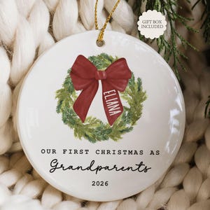 May include: A round white ceramic ornament featuring a green pine wreath illustration with a red bow. The bow has the name Eliana printed on it. Below the wreath, the text reads Our First Christmas As Grandparents 2026. A gold string is attached.