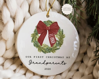 May include: A round white ceramic ornament featuring a green pine wreath illustration with a red bow. The bow has the name Eliana printed on it. Below the wreath, the text reads Our First Christmas As Grandparents 2026. A gold string is attached.