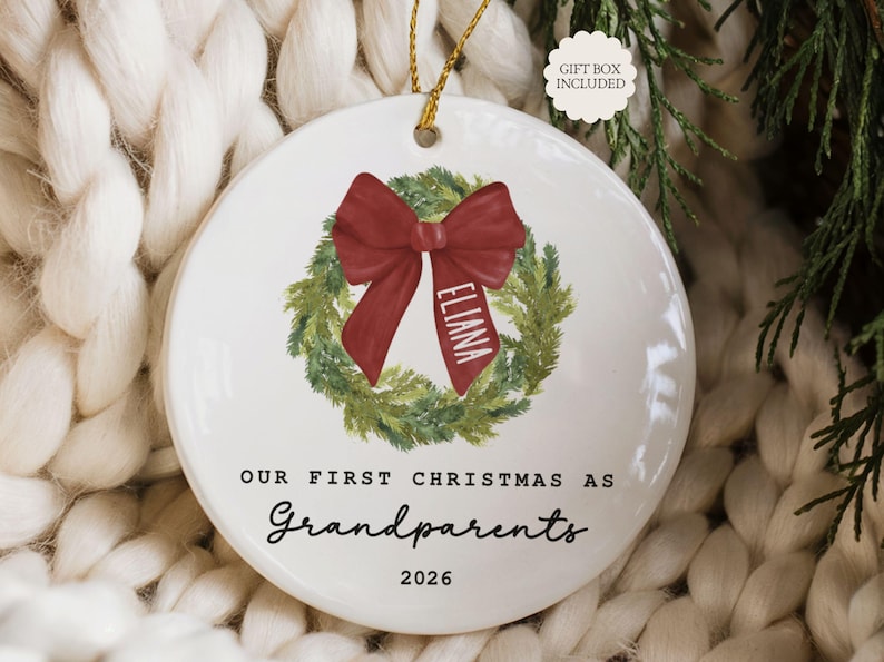 May include: A round white ceramic ornament featuring a green pine wreath illustration with a red bow. The bow has the name Eliana printed on it. Below the wreath, the text reads Our First Christmas As Grandparents 2026. A gold string is attached.