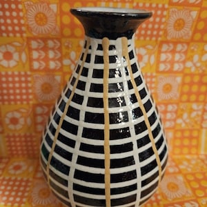 May include: A ceramic vase with a bulbous body and a flared rim, featuring a glossy black and white grid pattern overlaid with vertical tan drip lines that extend from the neck down the sides of the vessel, creating a distinct textured appearance.