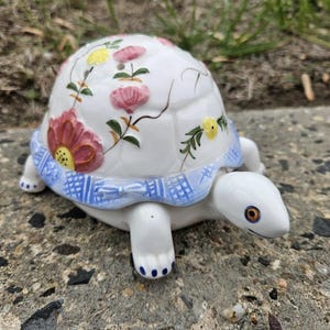 May include: A white ceramic turtle figurine with a floral design. The turtle's shell is decorated with pink and yellow flowers and green leaves. The turtle has a blue patterned band around its middle and measures approximately 5 inches long.