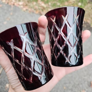 May include: Two ruby-red, cut-glass tumblers with a diamond pattern are held in a hand. The glasses have a slightly curved shape and reflect light, showcasing the intricate design. The background is blurred, suggesting an outdoor setting.