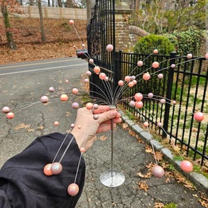May include: A decorative metal sculpture featuring a central vertical rod with a circular base and numerous thin, radiating wire arms. Each wire arm is tipped with a small, spherical bead in varying shades of pink, peach, and iridescent lavender.
