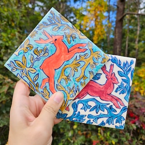 May include: Two ceramic tiles with a blue, white, and gold floral pattern. Each tile features a red deer in a leaping pose.