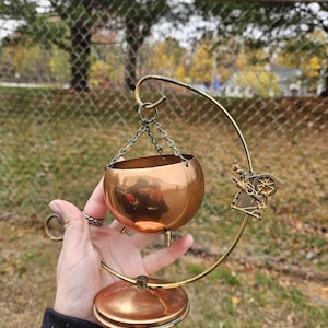 May include: A copper-colored incense burner with a round bowl suspended by chains from a curved brass stand. The stand features a decorative figure and a circular base. The burner is held by a person.