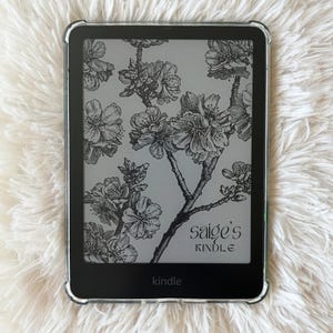 May include: A black Kindle e-reader with a clear protective case. The screen displays a black and white floral design with the text "saige's KINDLE" and "kindle". The e-reader is resting on a white, fluffy surface.