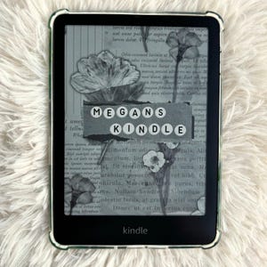 Puede incluir: Un lector de libros electrónicos Kindle negro con una funda protectora transparente. La pantalla muestra un diseño floral y el texto "MEGANS KINDLE". El dispositivo está sobre una superficie blanca texturizada.