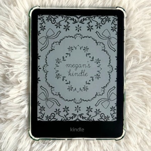 May include: A black Kindle e-reader with a clear protective case. The screen shows a floral design with the text "megan's kindle." The e-reader is on a white, fluffy surface.
