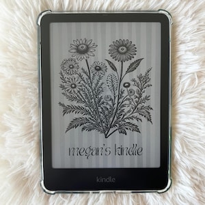 May include: A black Kindle e-reader with a clear protective case. The screen displays a floral illustration with the text "megan's kindle." The Kindle is on a white, fluffy surface.