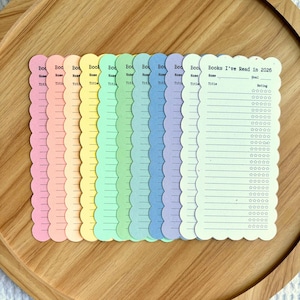 May include: A collection of pastel-colored book trackers with scalloped edges, arranged on a wooden tray. Each tracker has space for the book title, name, and a rating system. One tracker reads "Books I've Read in 2026."