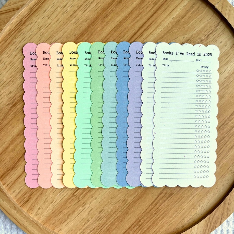 May include: A collection of pastel-colored book trackers with scalloped edges, arranged on a wooden tray. Each tracker has space for the book title, name, and a rating system. One tracker reads "Books I've Read in 2026."