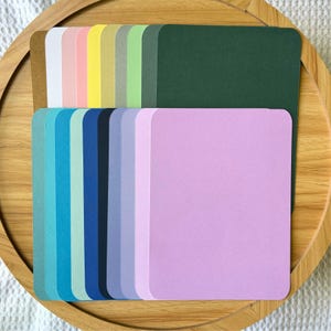 May include: A collection of colorful square cards in various shades, including pink, blue, green, and purple. The cards are arranged on a wooden tray, showcasing a range of pastel and vibrant hues. The cards have rounded corners.