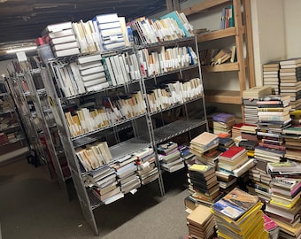 May include: A large collection of books organised on metal shelves and stacked on the floor. The books are mostly paperback and hardback novels, with some textbooks and other non-fiction books.