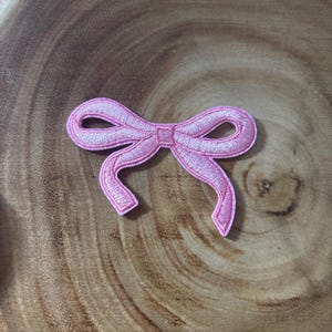 May include: A light pink embroidered patch in the shape of a bow with two loops and two trailing ribbon ends. The design features a dense satin stitch border and a textured thread fill, creating a defined outline and a soft, glossy appearance.