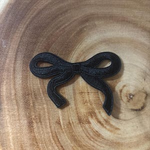 May include: A black embroidered patch in the shape of a bow. The patch features a textured surface with stitched detailing along the edges, forming two loops and two trailing ribbon ends that curve downwards in a symmetrical design.