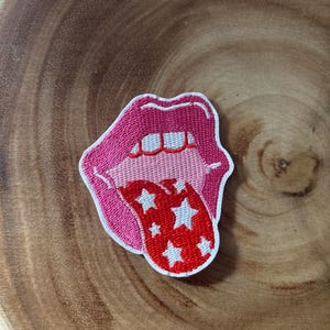 Puede incluir: Un parche bordado que muestra un par de labios abiertos con contornos rosas, dientes blancos y una lengua de color rojo brillante decorada con múltiples estrellas blancas. El parche tiene un borde blanco y sigue el contorno de la boca.