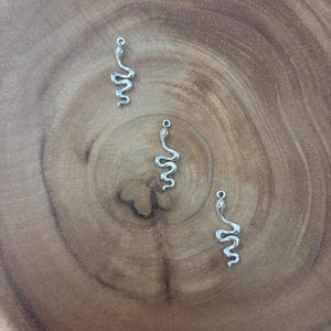 May include: Three silver-toned metal snake charms, each featuring a slender, winding body and a small loop at the top for attachment. The charms have a polished, reflective surface and are arranged in a scattered formation on a wooden surface.