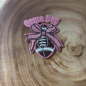 May include: An embroidered patch featuring a brown football with white laces, adorned with a pink bow and the text Game Day in pink lettering. The patch has a pink border and is designed for application to fabric surfaces as a decorative element.