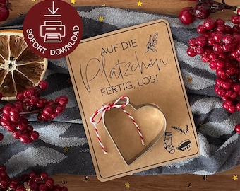 May include: A heart-shaped cookie cutter tied with red and white twine, resting on a brown card with German text. The card is decorated with small illustrations and sits on a patterned surface with berries and stars.